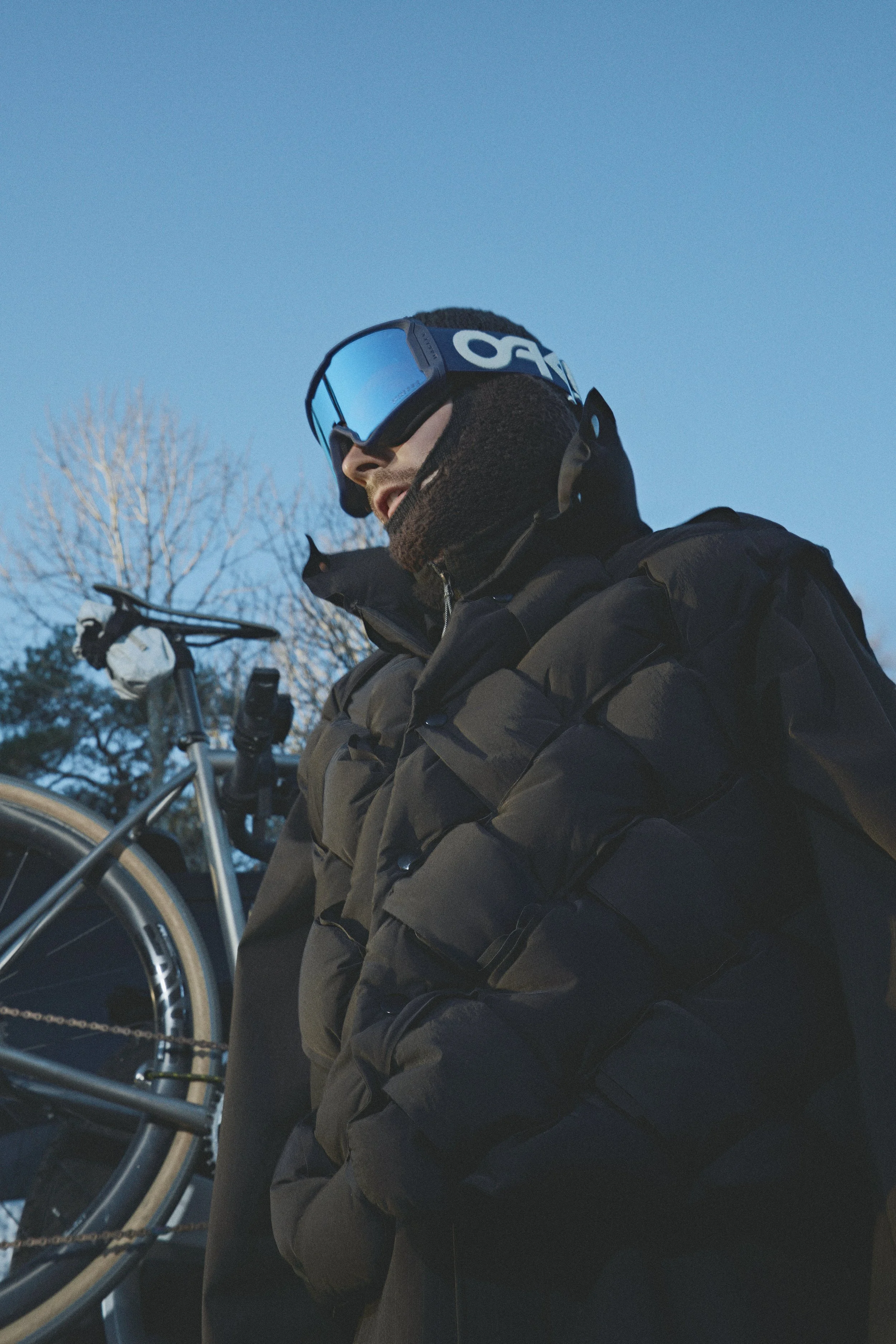 Patrick Stangby dressed in winter gear, wearing goggles, standing with a bicycle in an outdoor setting with leafless trees and a clear blue sky.