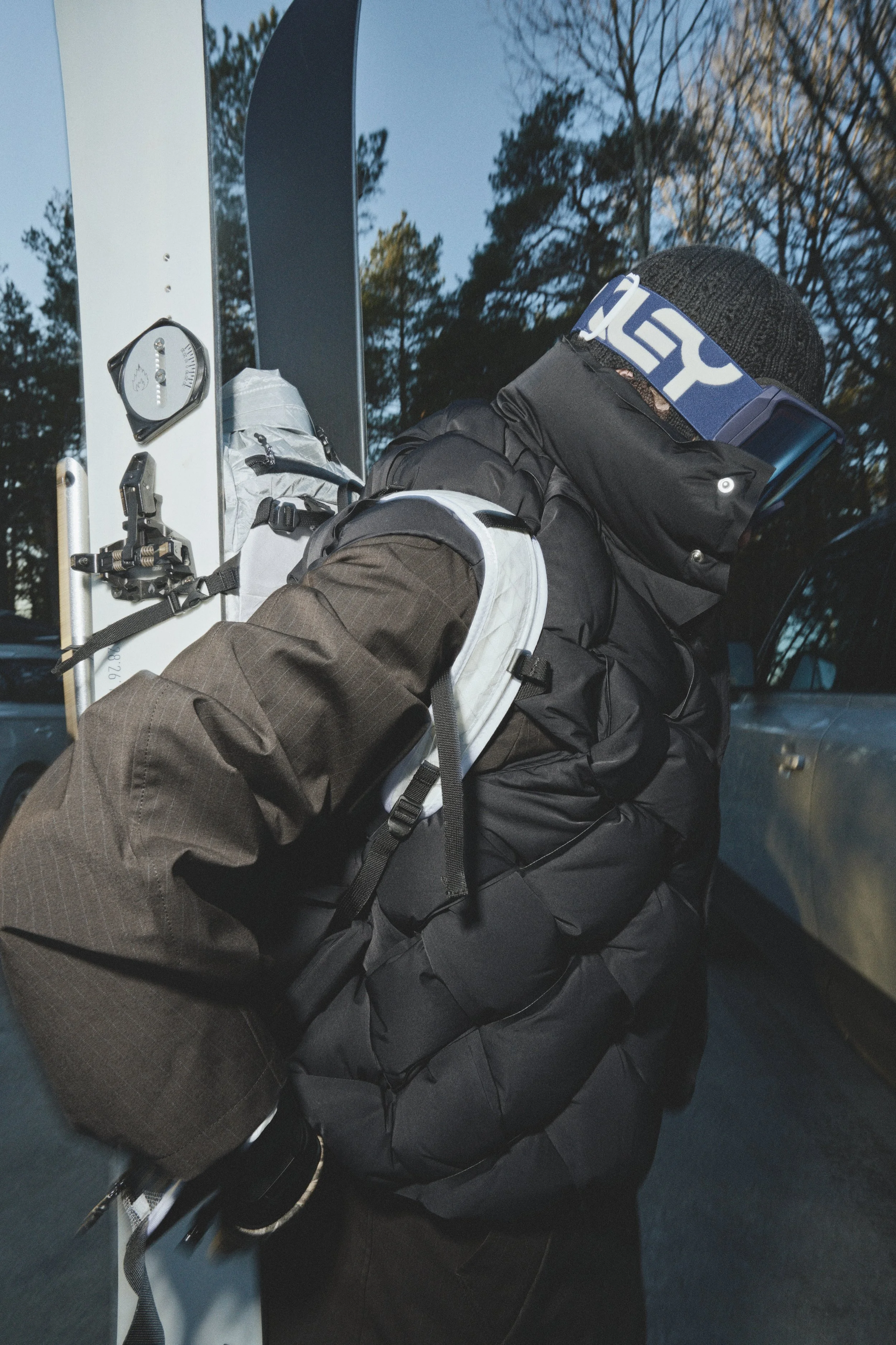 Patrick Stangby dressed for winter in black jacket, black gloves, and a black knit cap, with ski goggles and a headband. They are holding a pair of skis and a ski pole, standing outdoors next to a vehicle, with trees and a blue sky in the background.