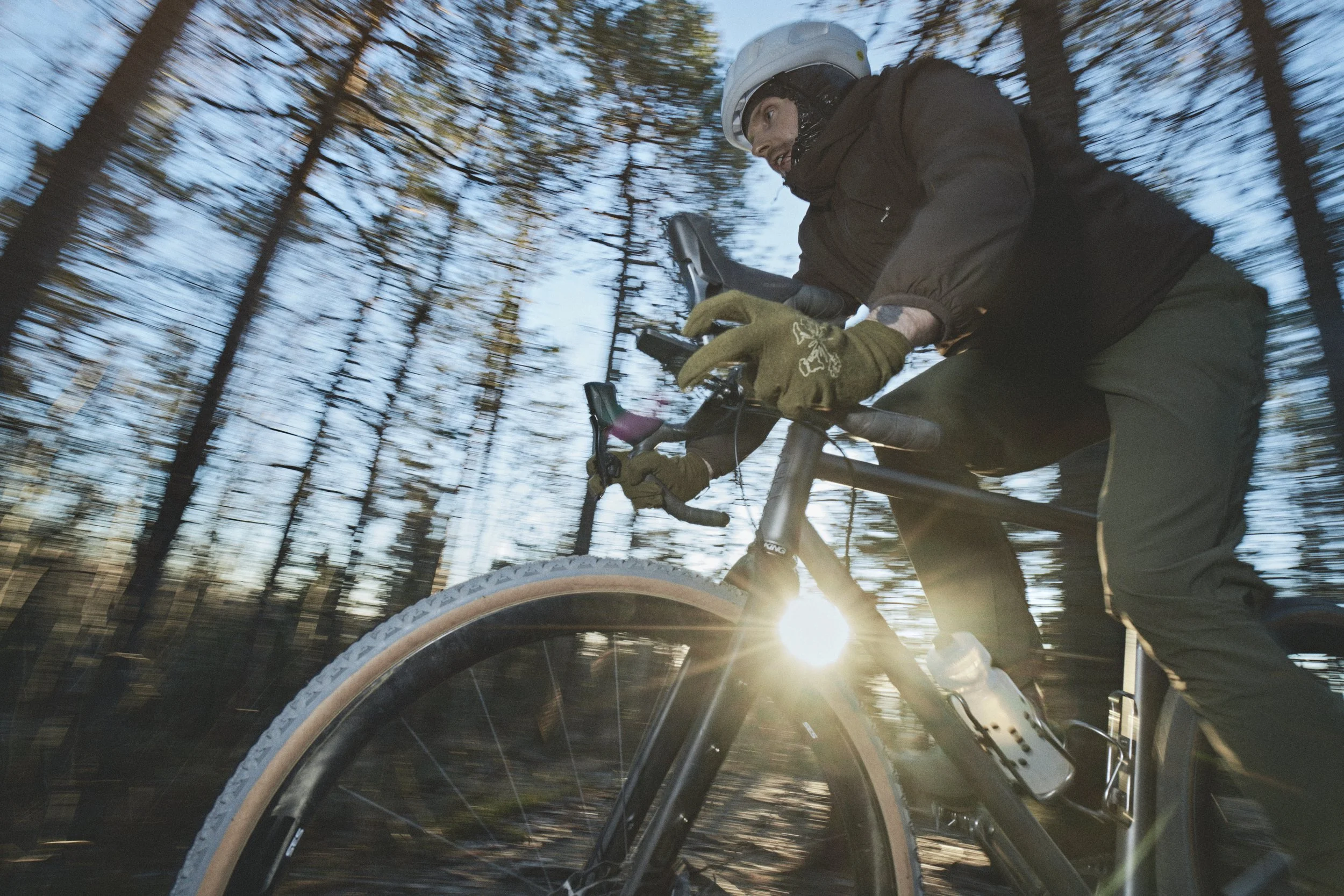 Patrick Stangby riding a mountain bike through a forest with tall trees, wearing a helmet and gloves, with sunlight peeking through the trees.