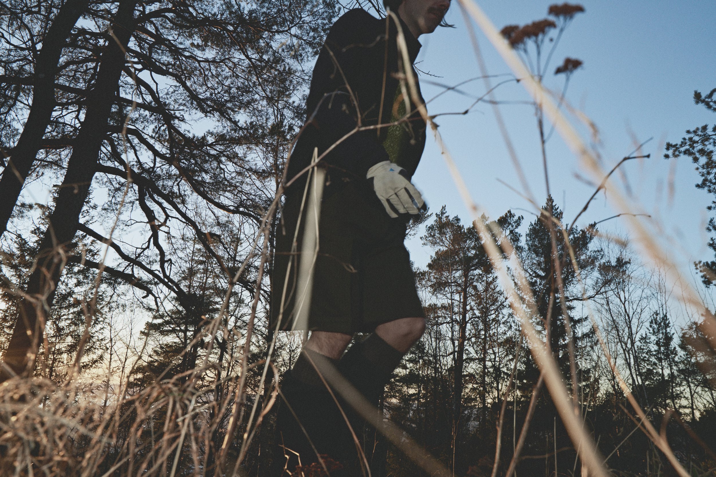 A man in black shorts, black jacket, and white gloves standing in a forest clearing at sunset, viewed from a low angle through dry grass and tree branches.