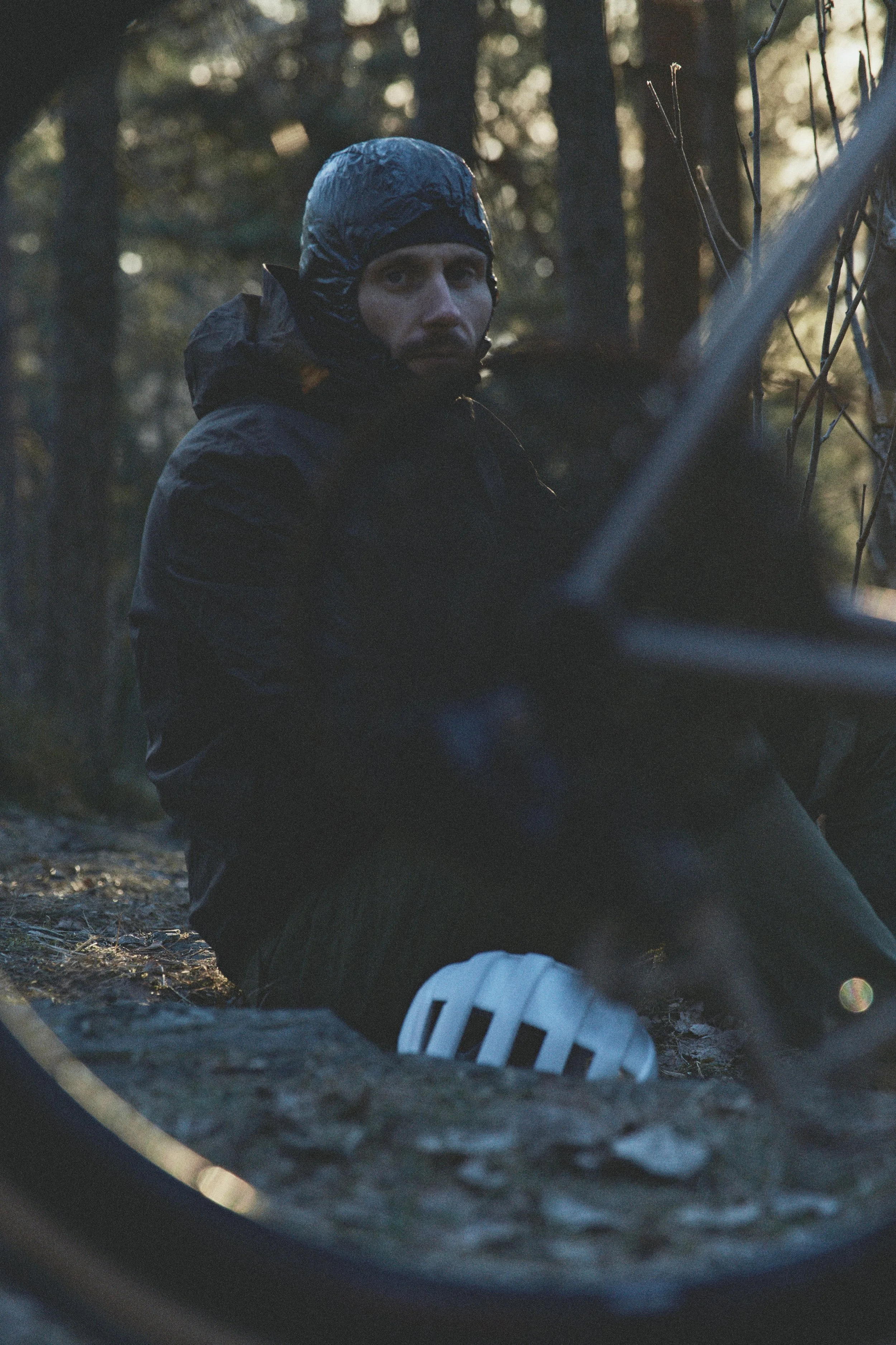 Patrick Stangby sitting on the ground in a forested area, dressed in black outdoor gear with a helmet, looking towards the camera with a serious expression, partially obscured by a bicycle frame.