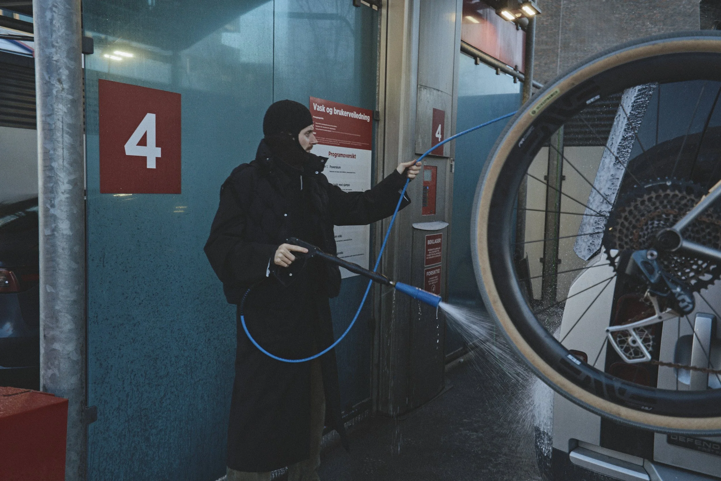 A person in black clothing and a beanie is pressure-washing a bicycle wheel outside. There is a blue wall and a red sign with the number 4 in the background.