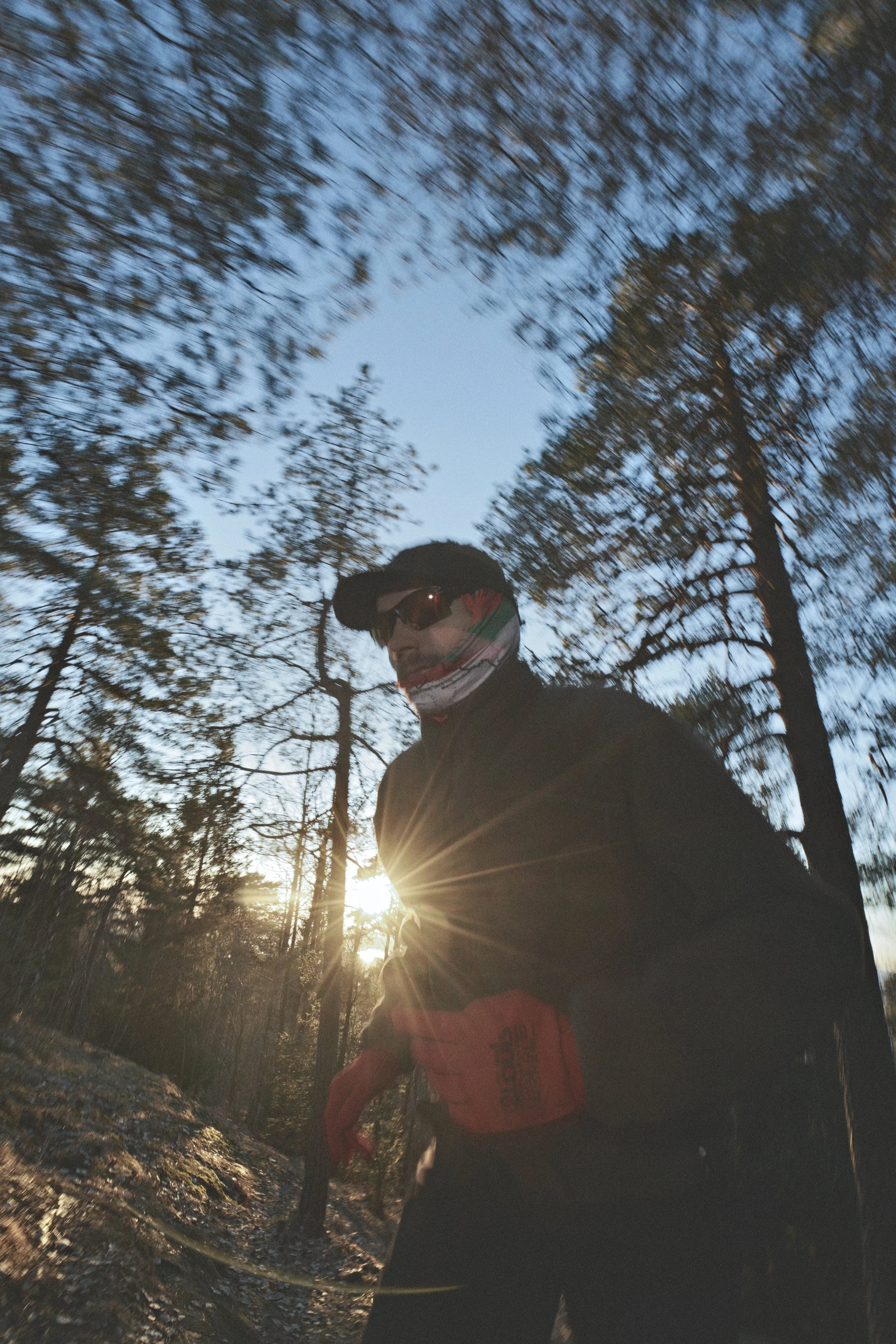 Patrick Stangby running in a forest during sunset, wearing a black jacket, red gloves, sunglasses, and a face covering, with sunlight shining through the trees.