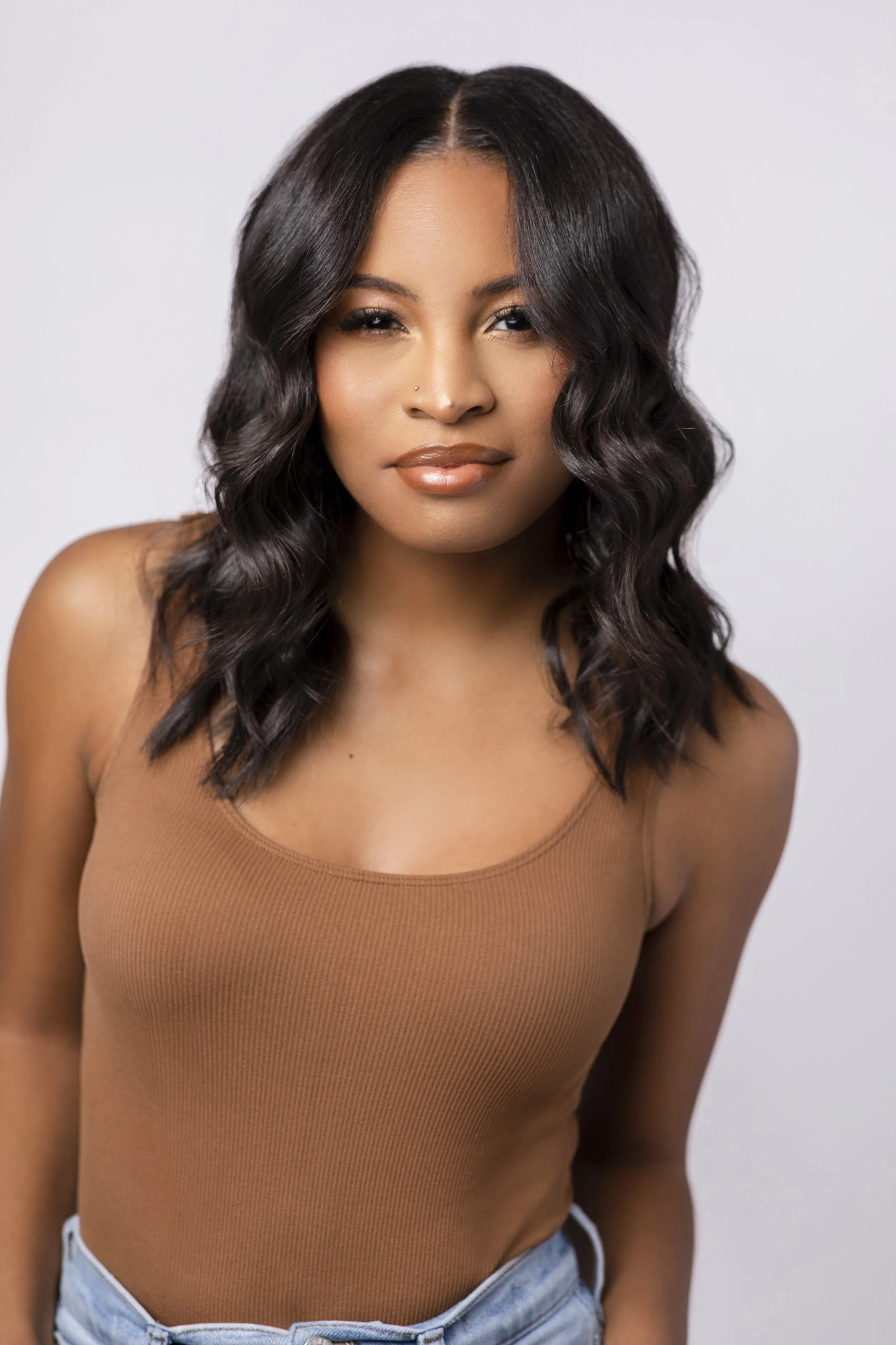 Close-up of an African American woman with wavy black hair, wearing a brown sleeveless top and light blue jeans, standing against a plain light-colored background.