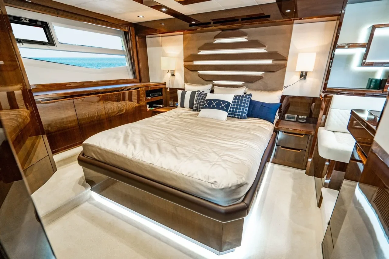 Luxury bedroom with a large bed, decorative pillows, and wooden accents. Ocean view window on the left, built-in side tables, and modern lighting fixtures.