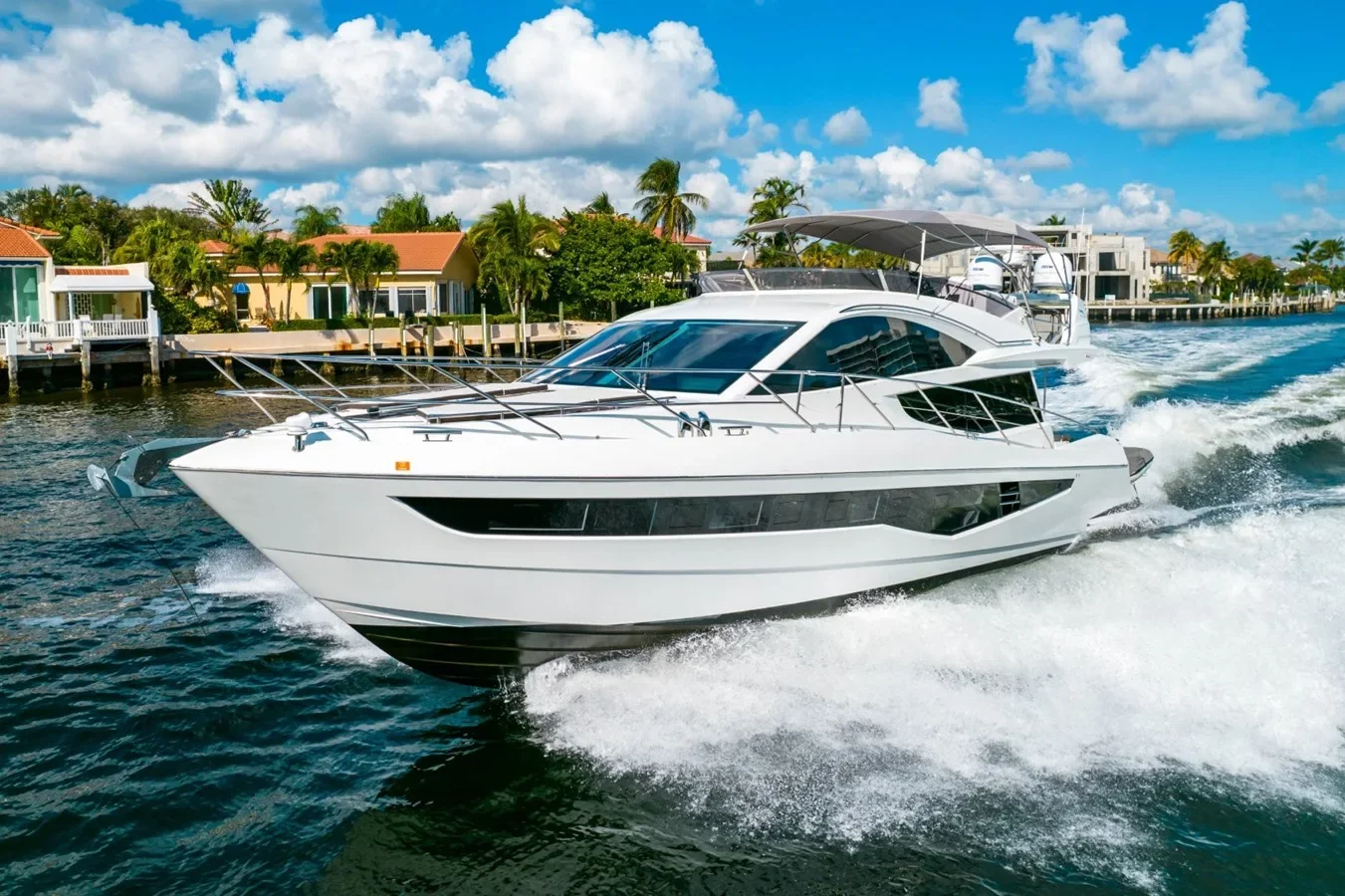 A white motor yacht speeding through the water with a shoreline of houses and palm trees in the background under a partly cloudy sky.