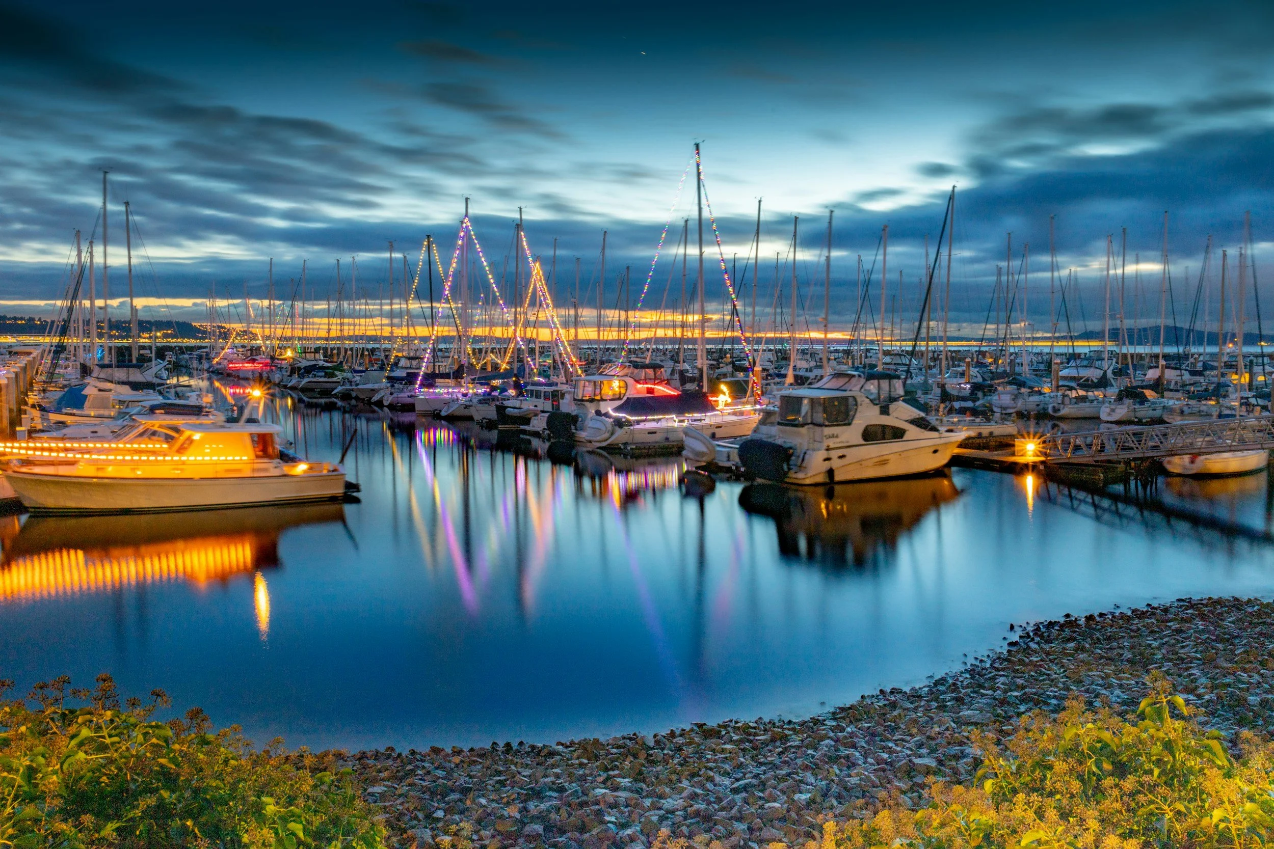 A marina at sunset with boats docked, string lights on boat masts, and a cloudy sky overhead.