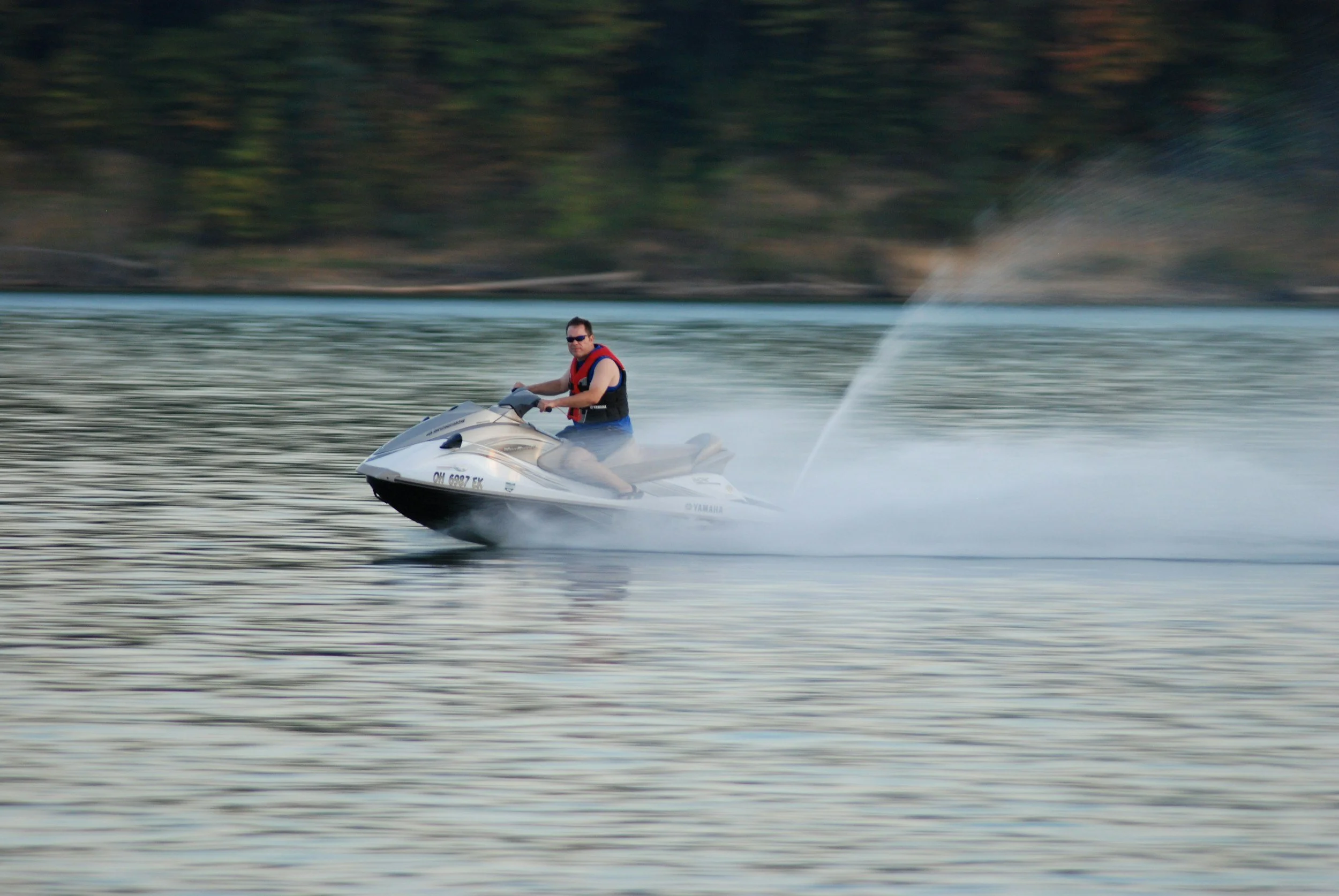 A man wearing sunglasses and a red life jacket riding a white jet ski on a body of water, with a forested shoreline in the background.