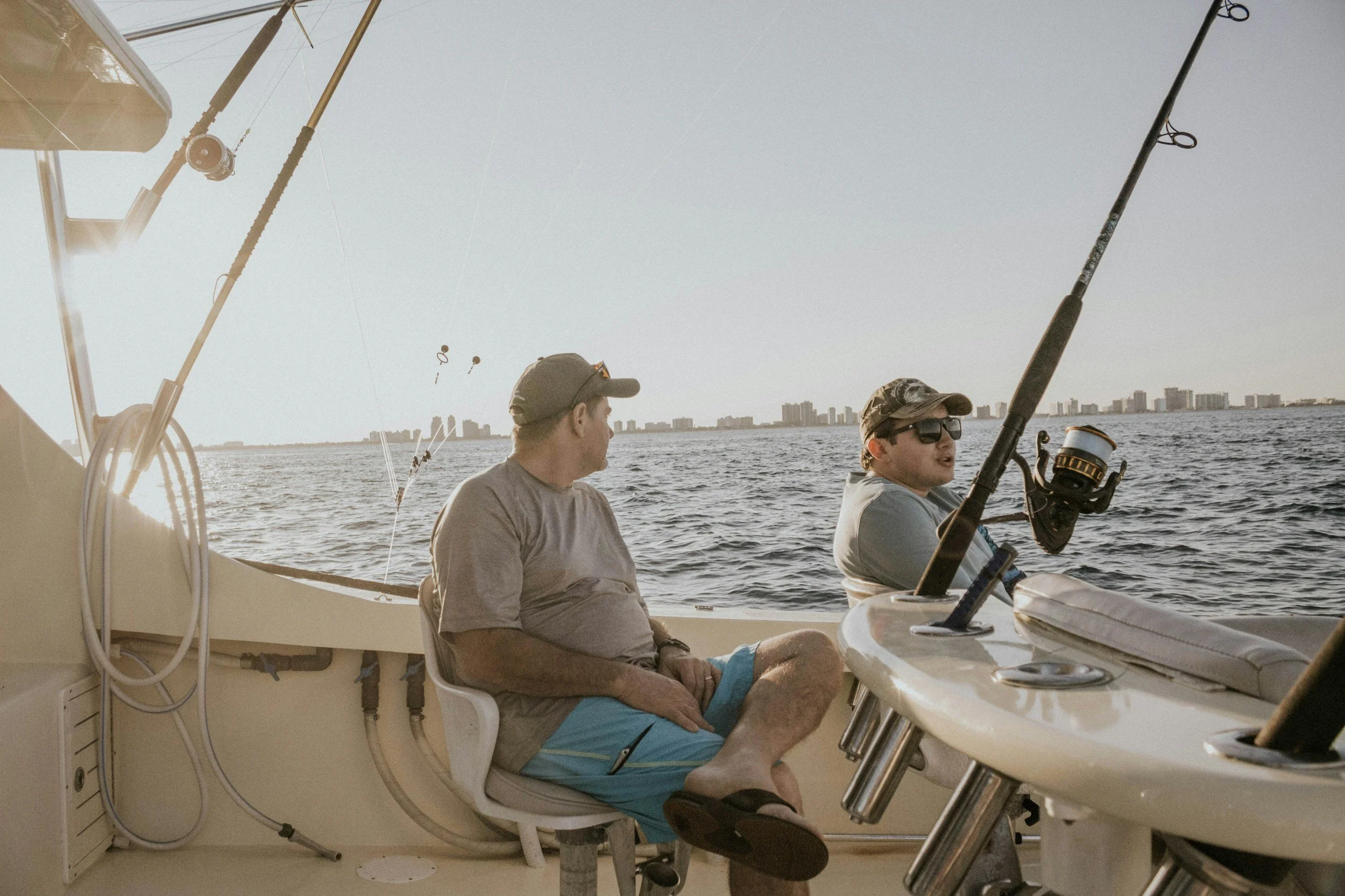 Two men sitting on a boat with fishing rods, overlooking a body of water with a city skyline in the background at sunset.