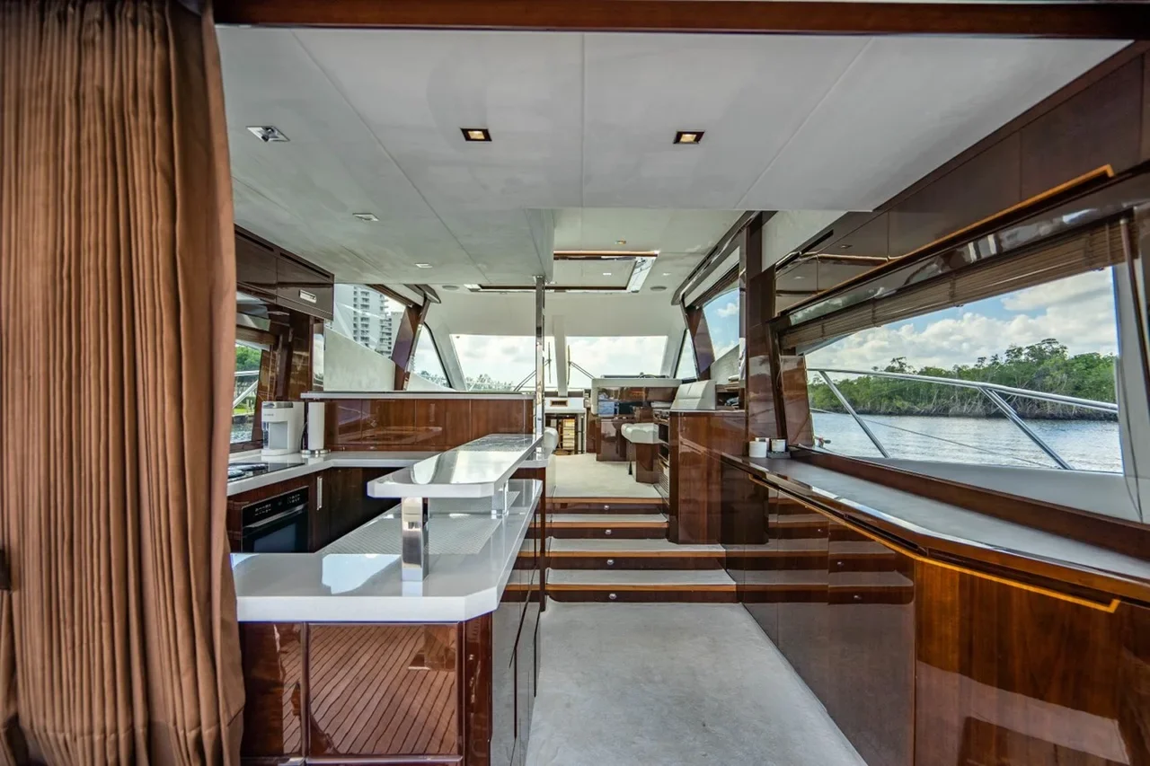 Interior of a yacht or boat with wood paneling, large windows showing water and greenery outside, a kitchen area with white countertops, and a staircase leading up to the helm station.