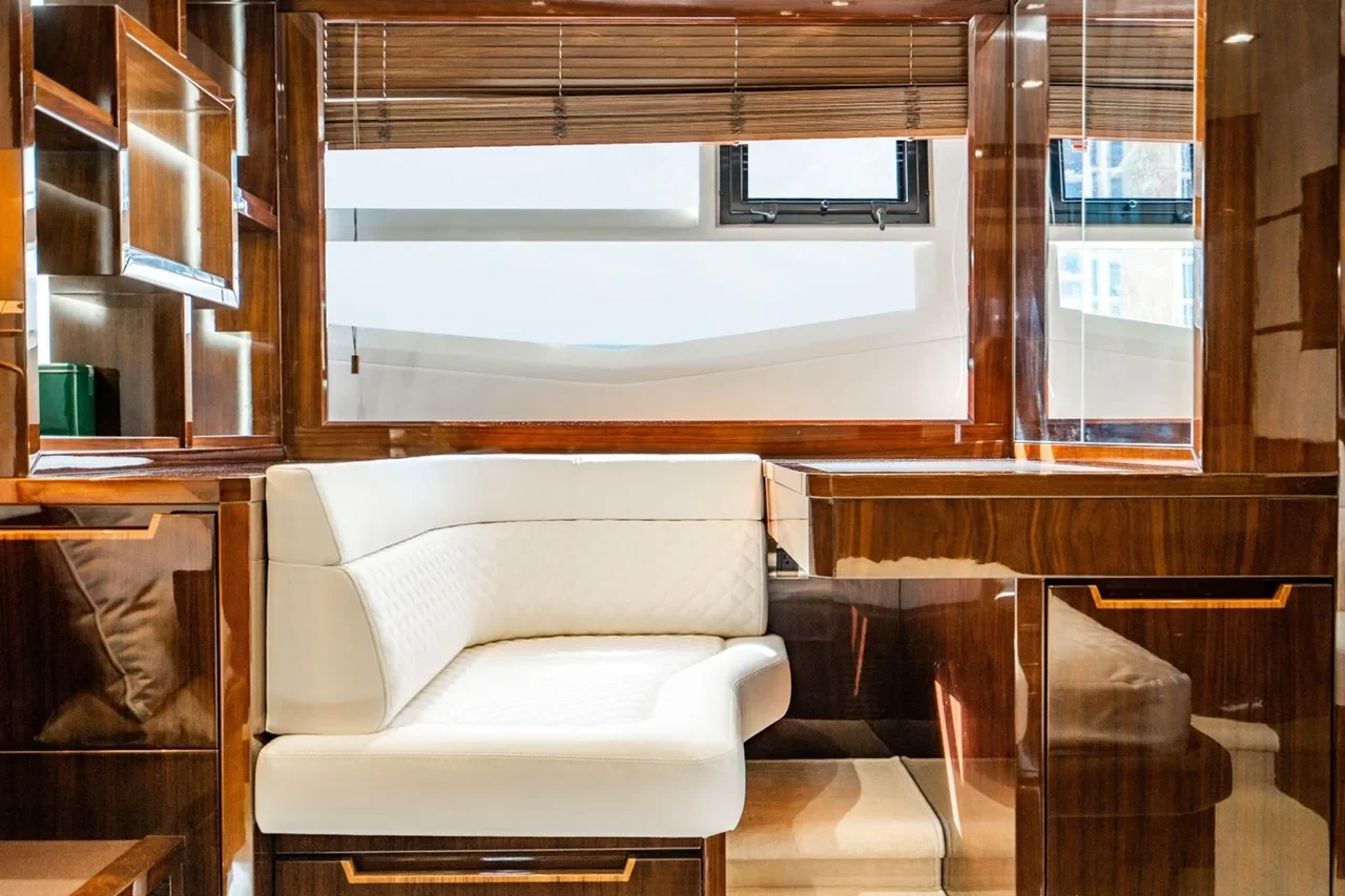 A white cushioned bench in a room with wooden built-in shelves and cabinets, a large mirror on the wall, a window with horizontal blinds, and reflections of the room's interior.