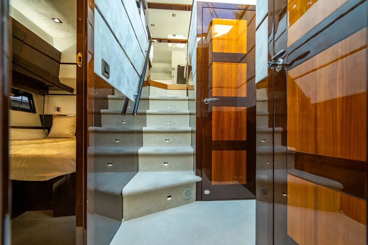 View of a luxurious yacht interior with a carpeted staircase, wooden paneling, glass partitions, and a glimpse of a bed in a cozy cabin.