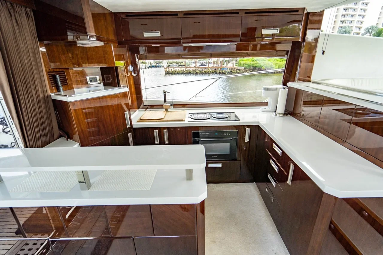 Interior view of a modern kitchen with dark wood cabinets, white countertops, and a large window overlooking a waterway with boats and buildings in the distance.