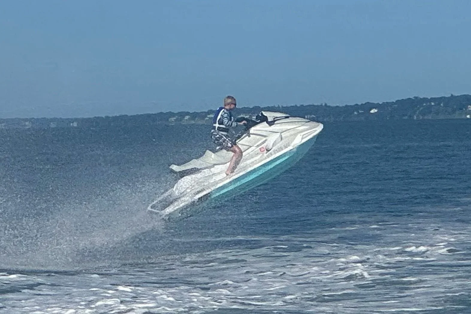 A person riding a white jet ski on the water, with a landmass and houses visible in the background.