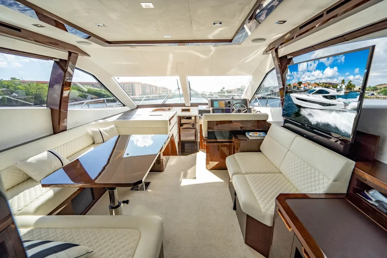 Interior of a luxury yacht with cream-colored leather seating, a wooden dining table, and a large flat-screen TV showing a yacht at sea, with windows revealing a marina outside.