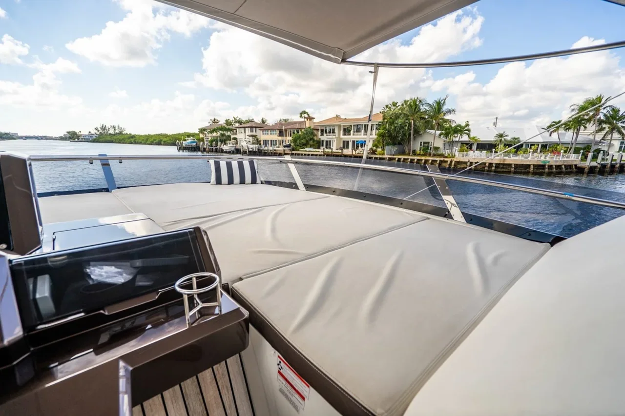 View from a luxury yacht showing a cushioned seating area with a striped pillow, a control panel, and a clear canopy. The waterway, waterfront houses, and palm trees are visible in the background under a partly cloudy sky.