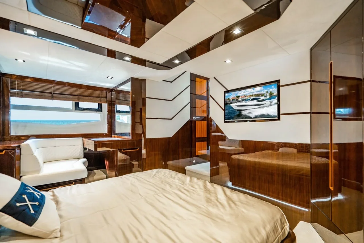 Luxury bedroom with wood paneling and a flat-screen TV showing a yacht, a bed with a nautical-themed pillow, and windows overlooking the ocean.