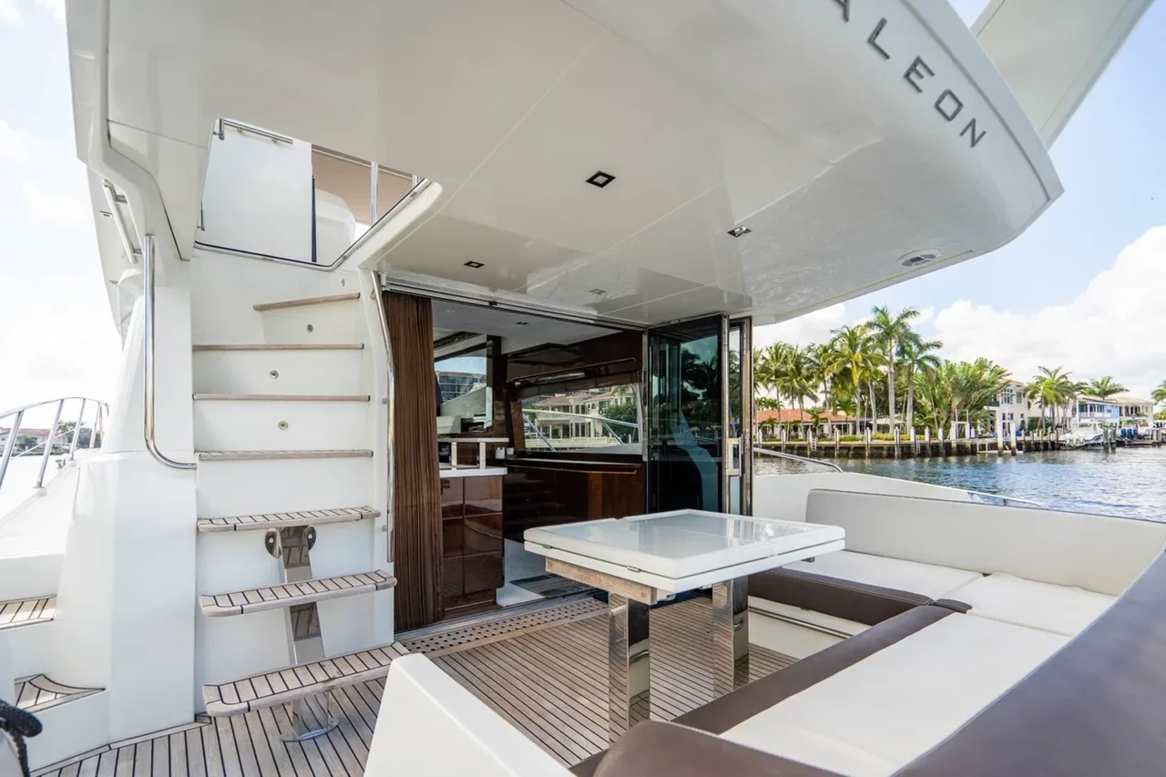 Upper deck of a yacht with seating area, a table, and a staircase, overlooking a waterway with palm trees and houses.