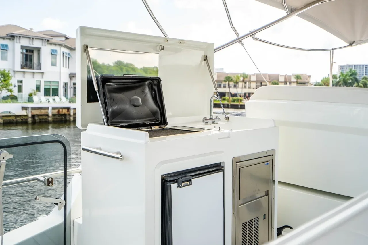 A small boat's outdoor kitchenette with a built-in grill and mini refrigerator, overlooking a waterway with residential buildings in the background.