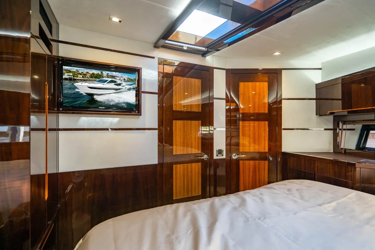 Interior of a yacht cabin with wood-paneled walls, a bed with white bedding, a wall-mounted TV showing a yacht on water, and small windows.