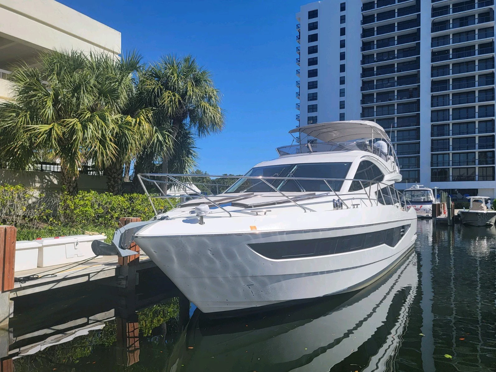 Luxury white yacht docked at a marina with high-rise buildings and palm trees in the background under a clear blue sky.