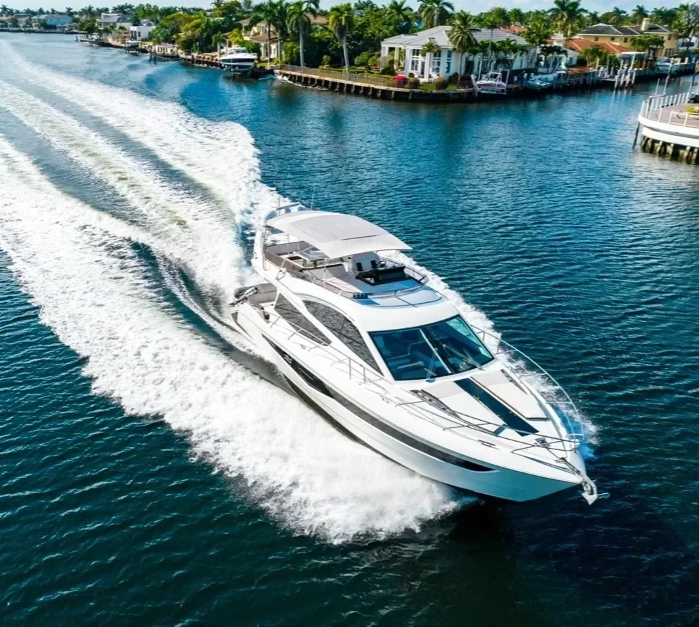 A white yacht moving through a canal with homes on the waterfront, palm trees, and blue skies in the background.