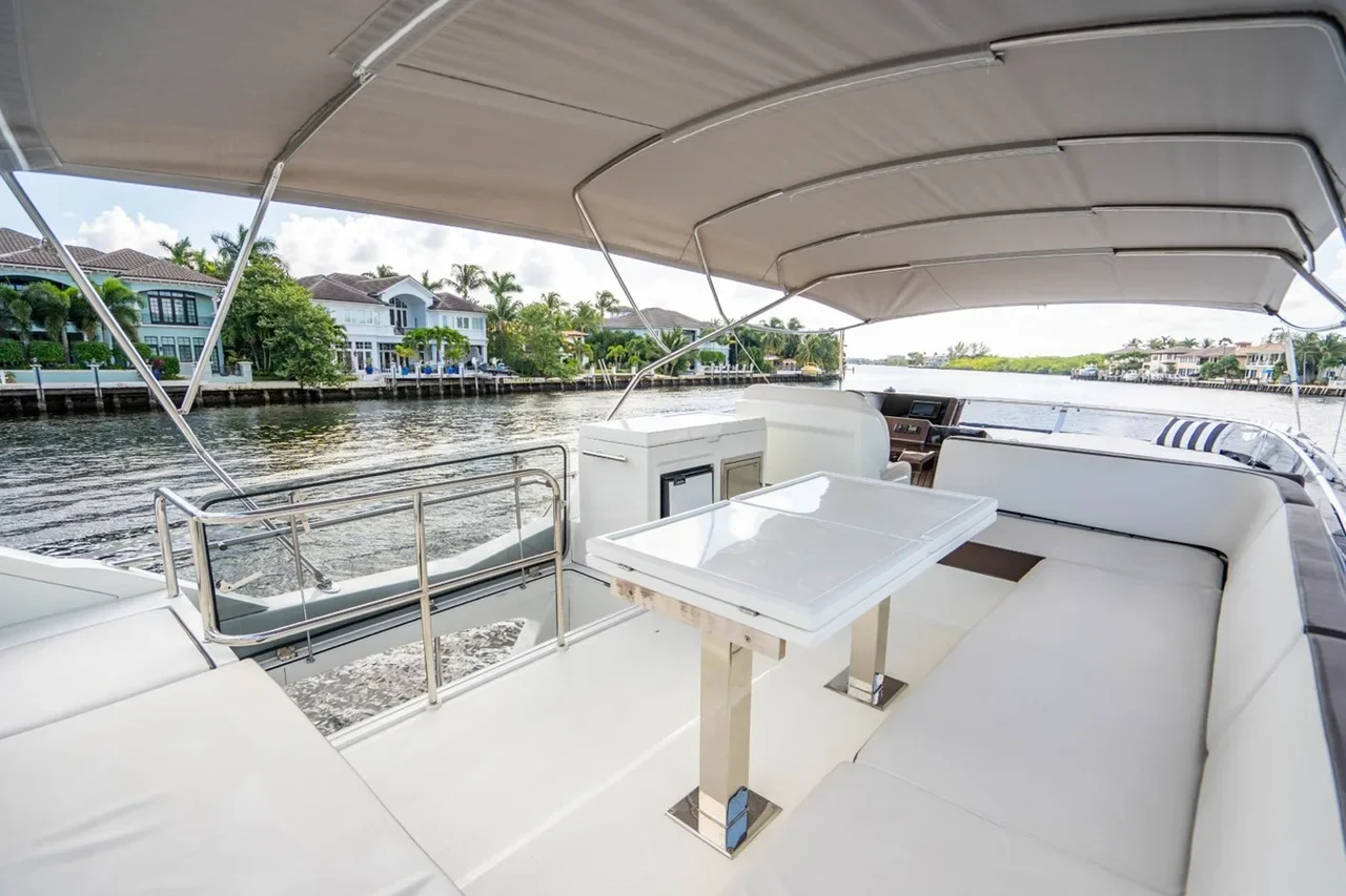 Upper deck of a luxury yacht with a white table, seating, and a canopy, overlooking water with waterfront houses and palm trees.