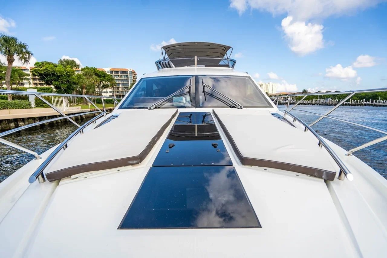 Front view of a white yacht docked in a marina with buildings and palm trees in the background under a partly cloudy sky.
