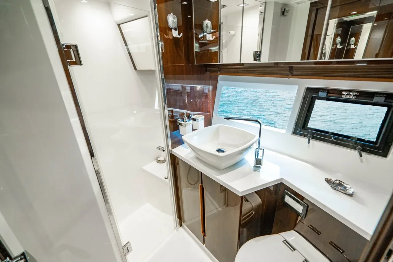 Compact bathroom on a boat with a white sink, a mirror, a small window showing water, a toilet, and wooden cabinets.