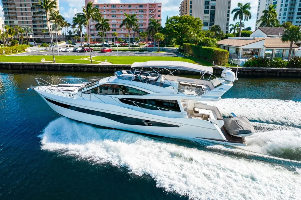 Two luxury yachts cruising in a waterway near a waterfront residential area with palm trees and high-rise buildings.