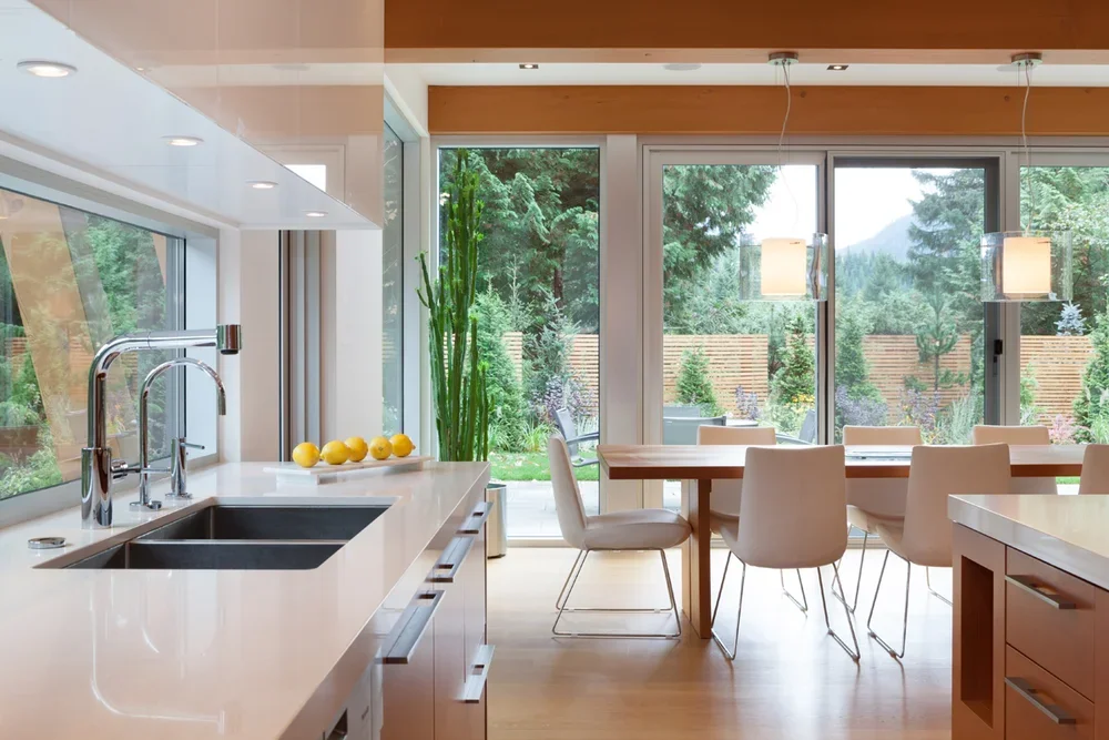 Modern kitchen with white countertops, stainless steel sink, large glass sliding doors, and a dining area with a wooden table and white chairs, overlooking a green garden.