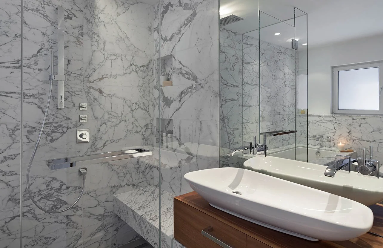 Modern bathroom with white marble walls, a bathtub with a sleek design, and a glass-enclosed shower area with marble accents. A window provides natural light.