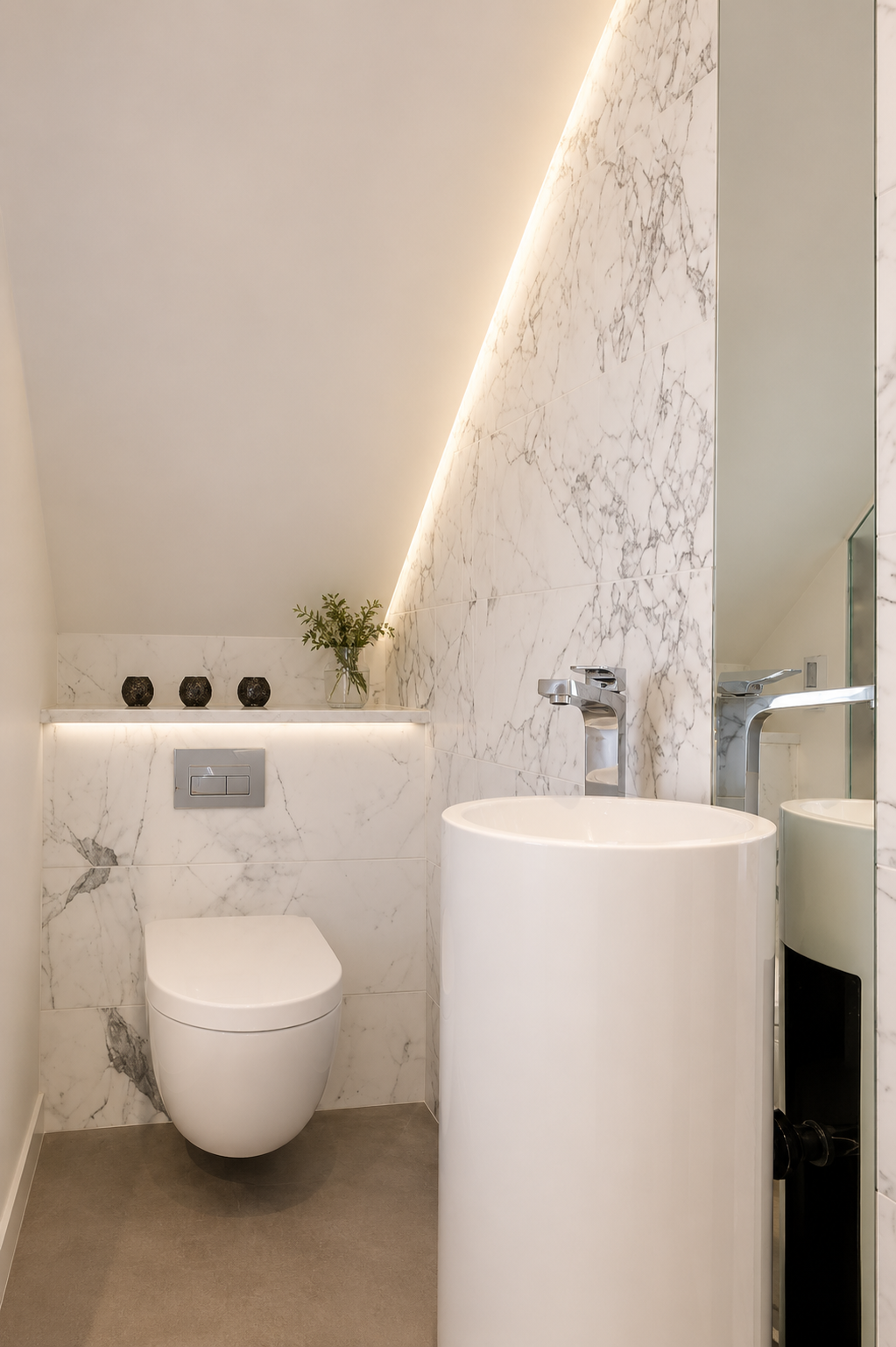 A modern bathroom with white marble walls, a floating toilet, a cylindrical sink, a large mirror, a small shelf with three black decorative objects and a small plant, and minimal lighting.