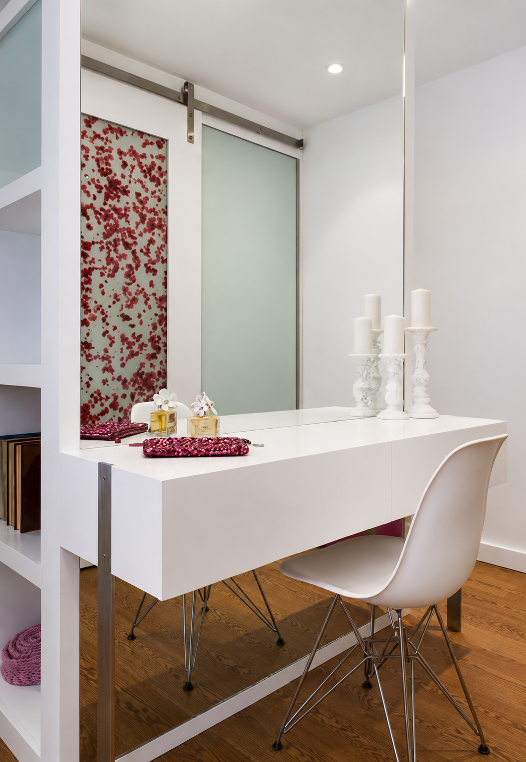 A modern white vanity table with decorative candles and perfume bottles, situated in a minimalistic room with white walls and a mirror. The room has a wooden floor and a white chair with metal legs.