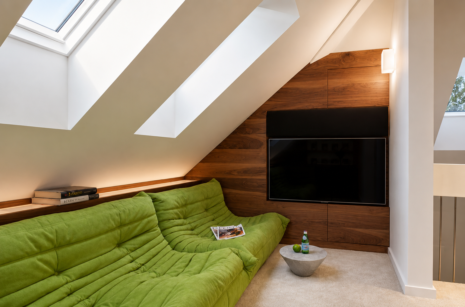 Living room with green sofa, wooden wall panel, flat-screen TV, skylight, books, and a small table with a water bottle, apple, and magazine.