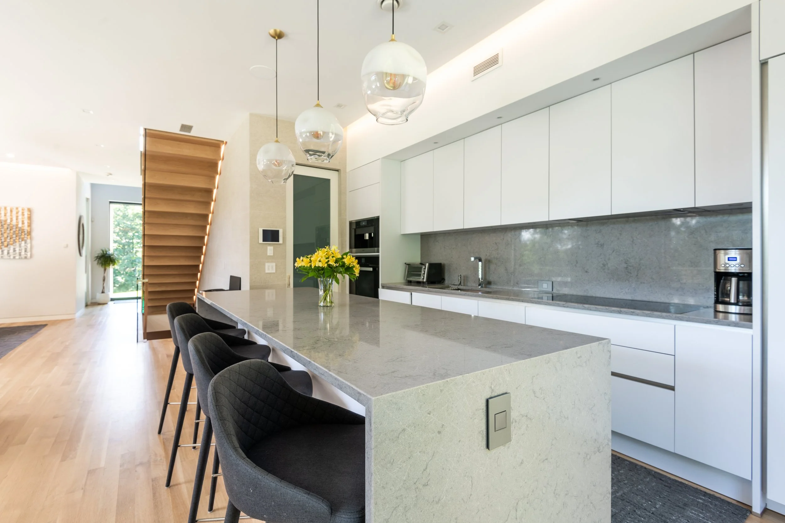 Modern kitchen with white cabinetry, marble countertops, hanging glass pendant lights, and a breakfast bar with black cushioned chairs. There's a vase of yellow flowers on the counter, a coffee maker, and a staircase with wooden steps and under-step lighting.