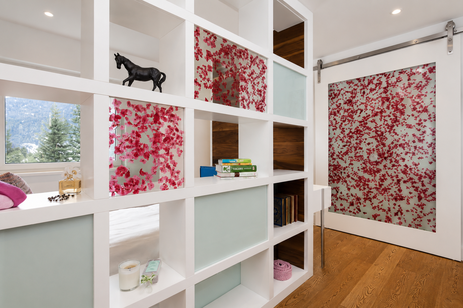 Interior of a room with a white shelving unit, decorative pink flower panels, a black horse figurine, books, a bottle of perfume, and a candle, with a window showing trees outside and a sliding door with a red floral pattern.