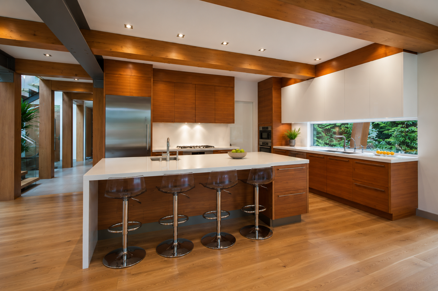 Modern kitchen with wooden cabinets, white countertops, stainless steel appliances, bar stools, and a large window overlooking greenery.
