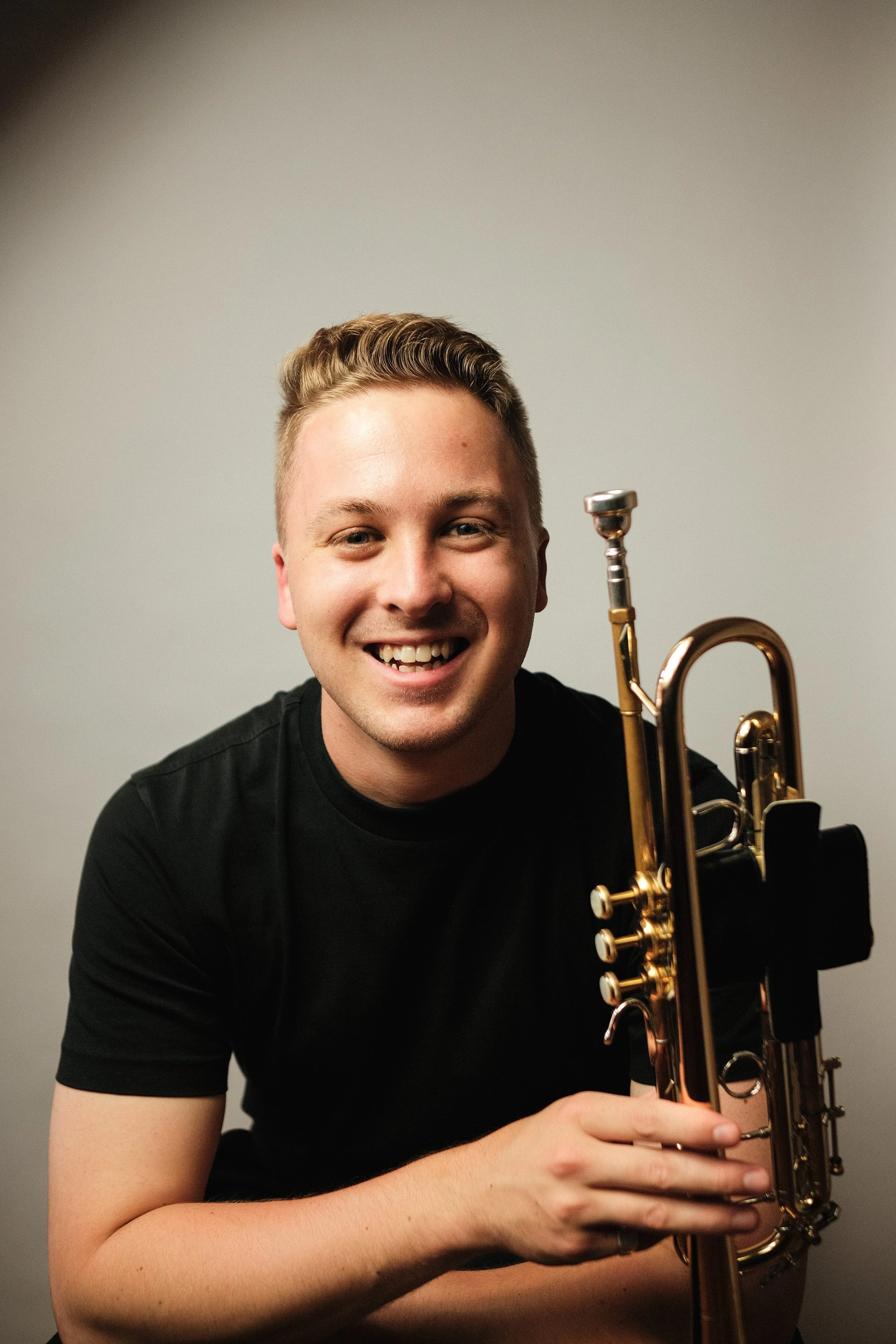 Young man with short blond hair smiling while holding a brass trumpet, wearing a black t-shirt, against a plain light grey background.
