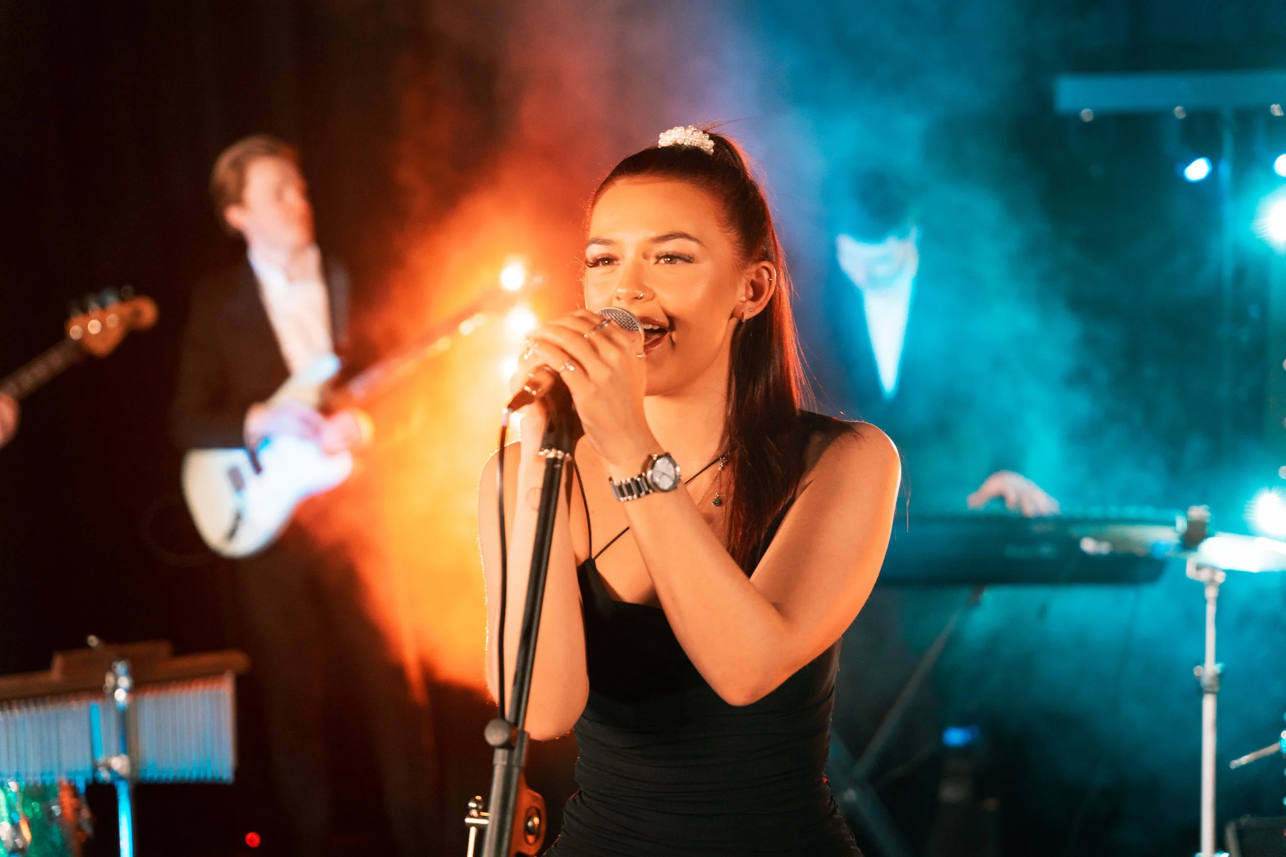 A young woman with dark hair in a high ponytail singing into a microphone on stage, with colorful stage lights and a blurred male guitarist in the background.