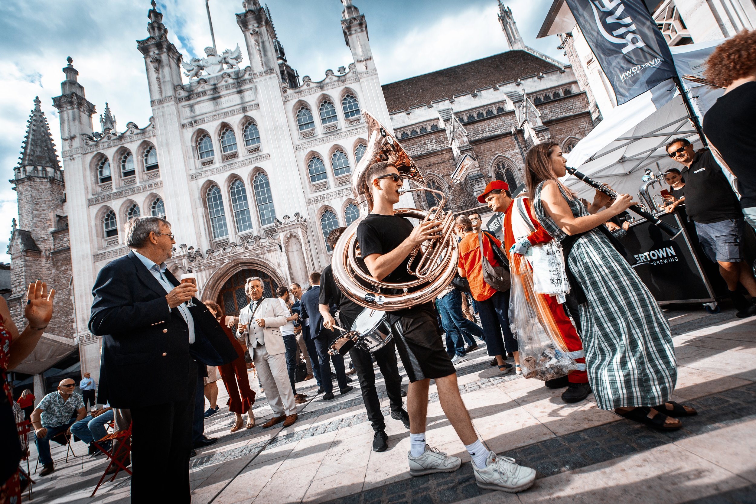 Street scene with musicians playing instruments, including a sousaphone and clarinet, in front of a historic Gothic-style building.