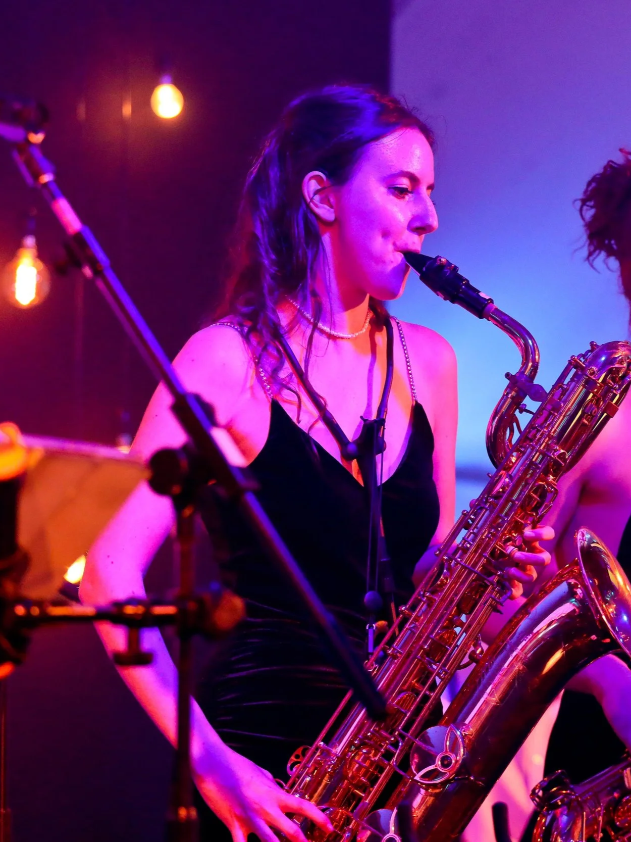 A woman playing a saxophone during a performance in a dimly lit venue with purple and pink lighting.