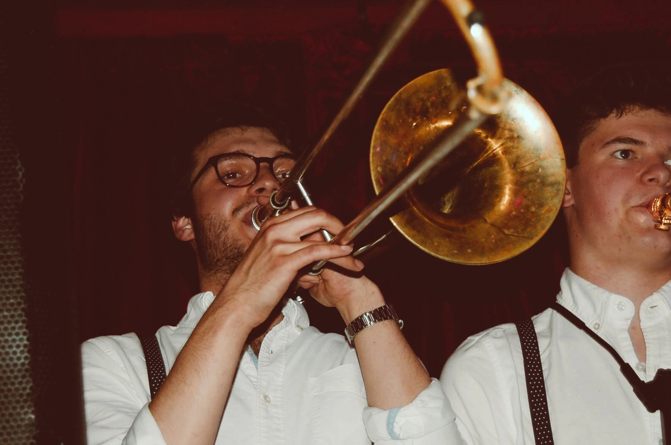 Two men wearing white shirts and suspenders playing a trombone together in a dimly lit room.