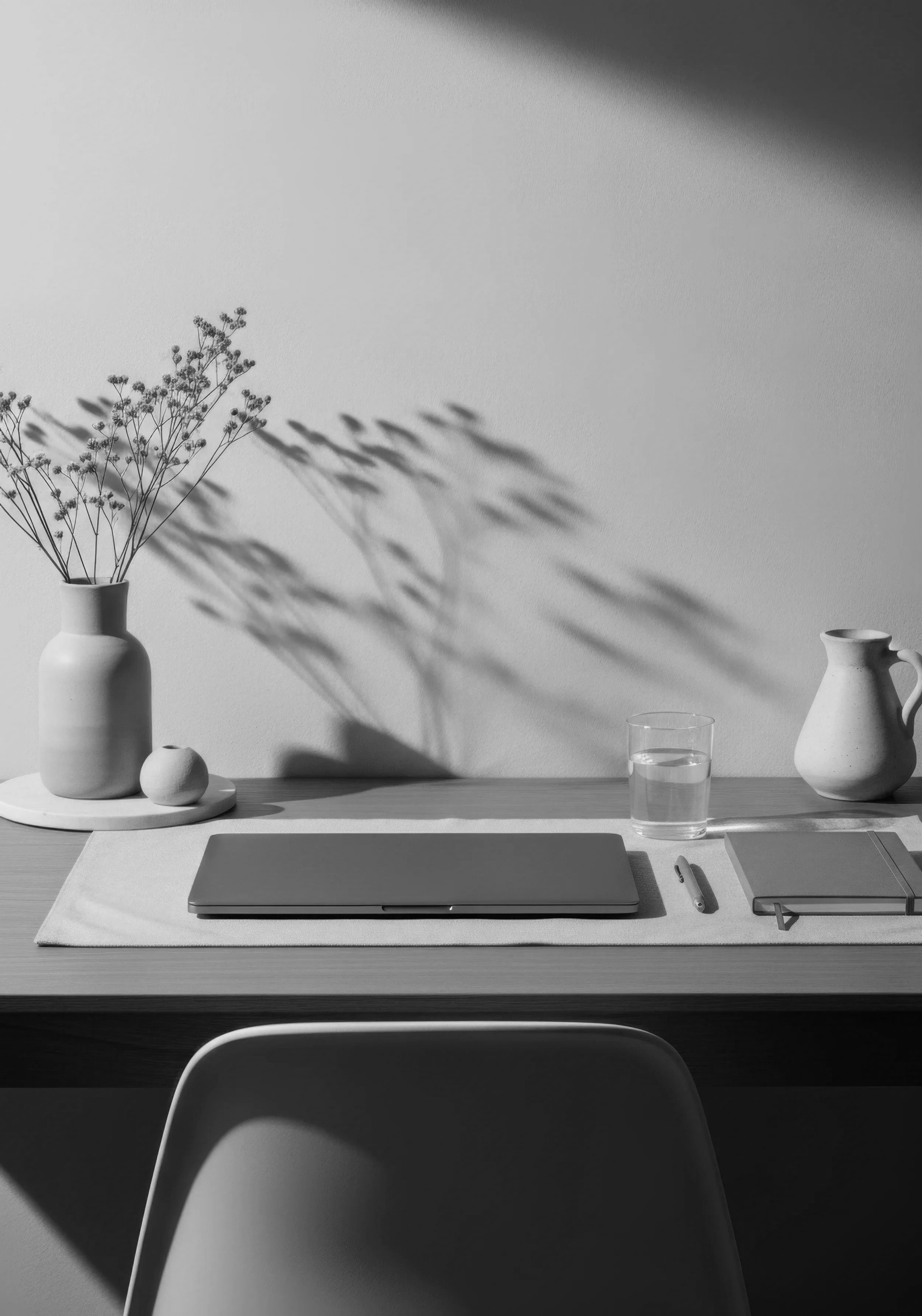 Black and white photo of a minimalist workspace with a laptop, a closed notebook, a pen, a glass of water, and decorative vases with dried flowers on a wooden table, creating a calm and organized atmosphere.