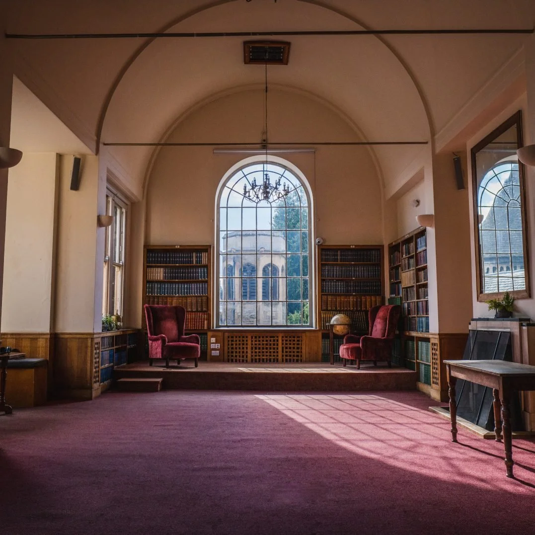A posh Cambridge meeting room with a large arched window, red velvet chairs, wooden bookshelves filled with books, and a chandelier hanging from the ceiling.