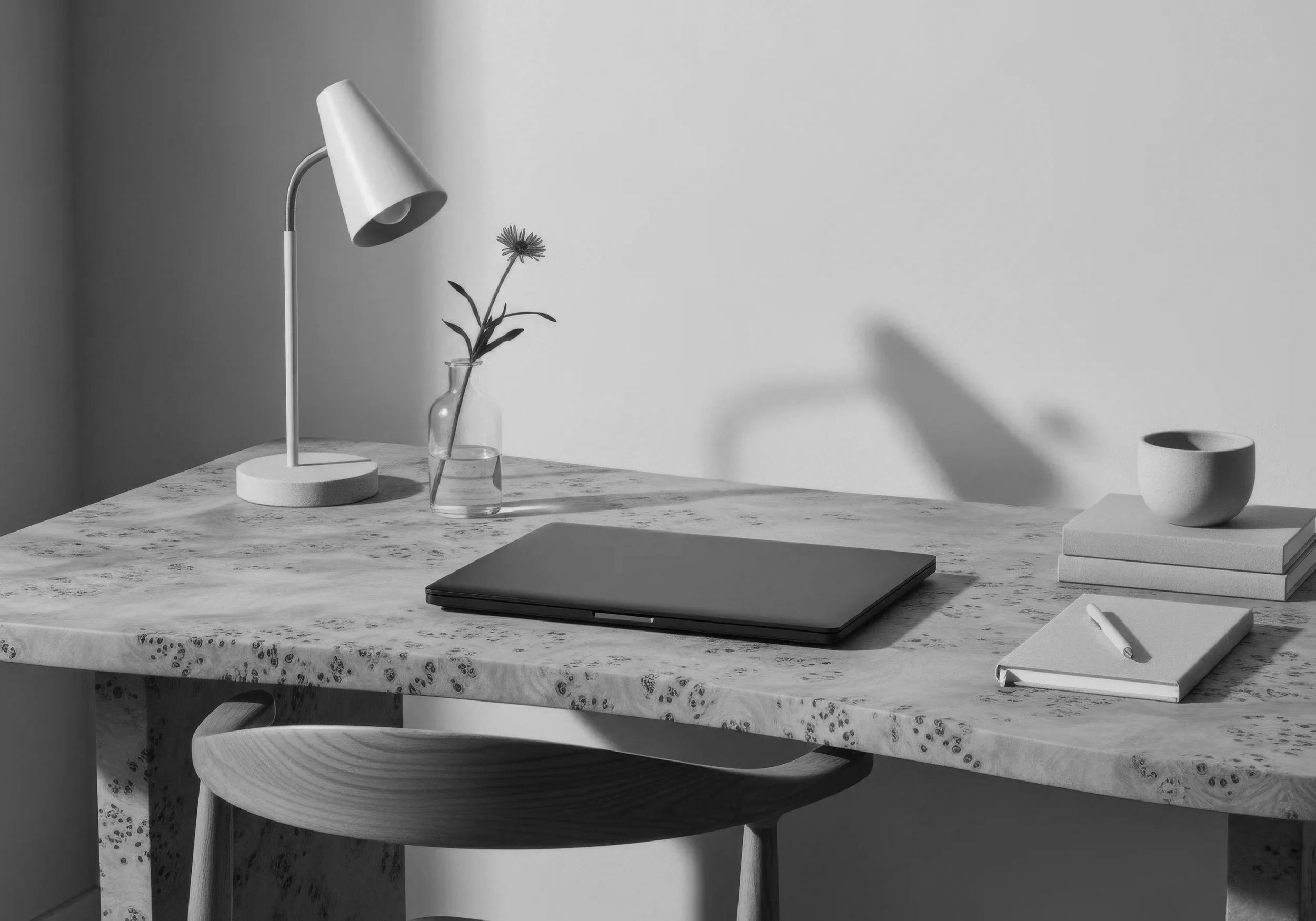 A black-and-white photo of a minimalist workspace with a marble desk, a closed laptop, a stack of notebooks, a small vase with a flower, and a modern lamp.