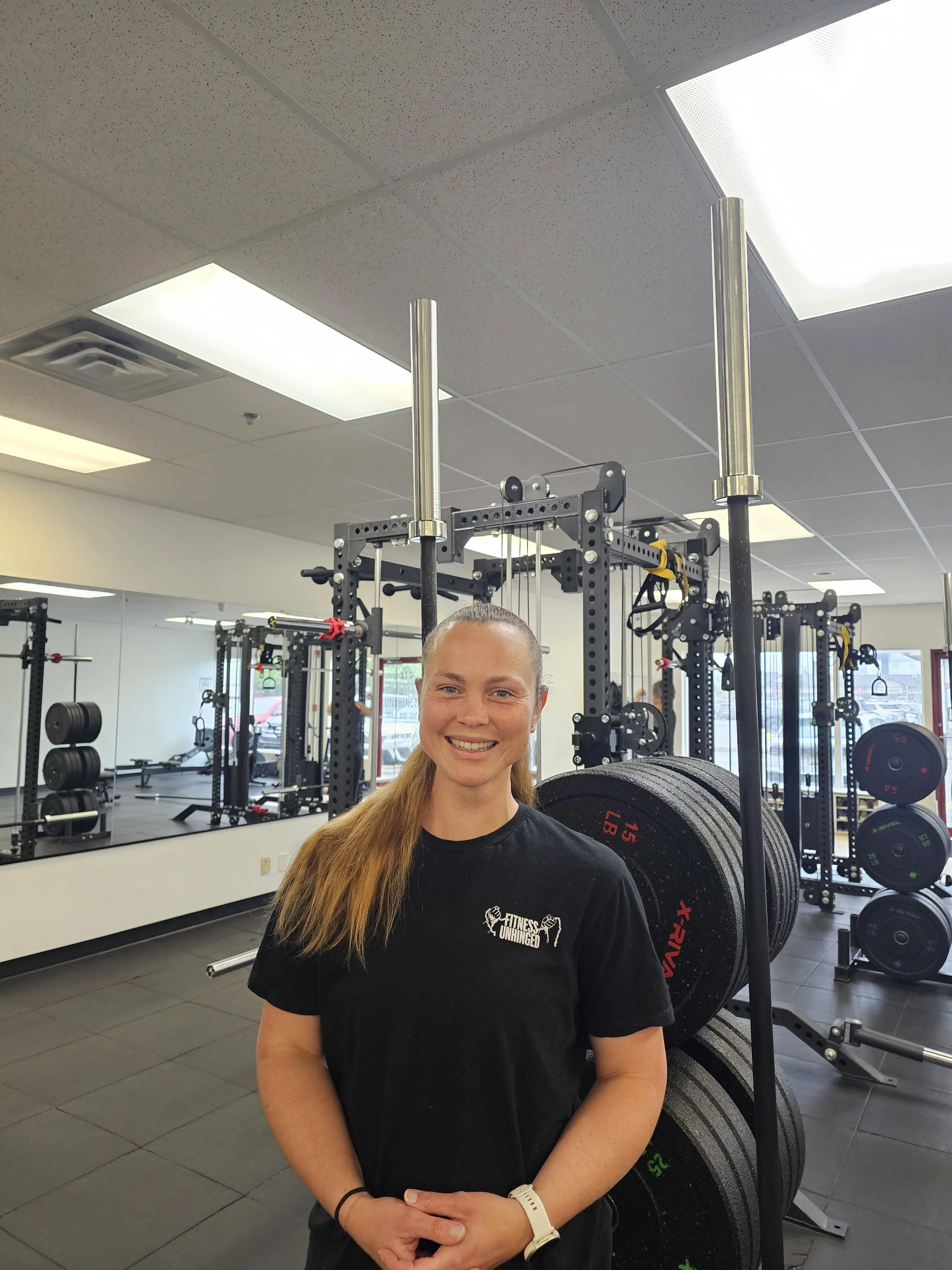 A smiling woman with long hair tied back, wearing a black T-shirt with the text 'Fitness Unhinged', standing in a gym surrounded by weightlifting equipment and mirrors.