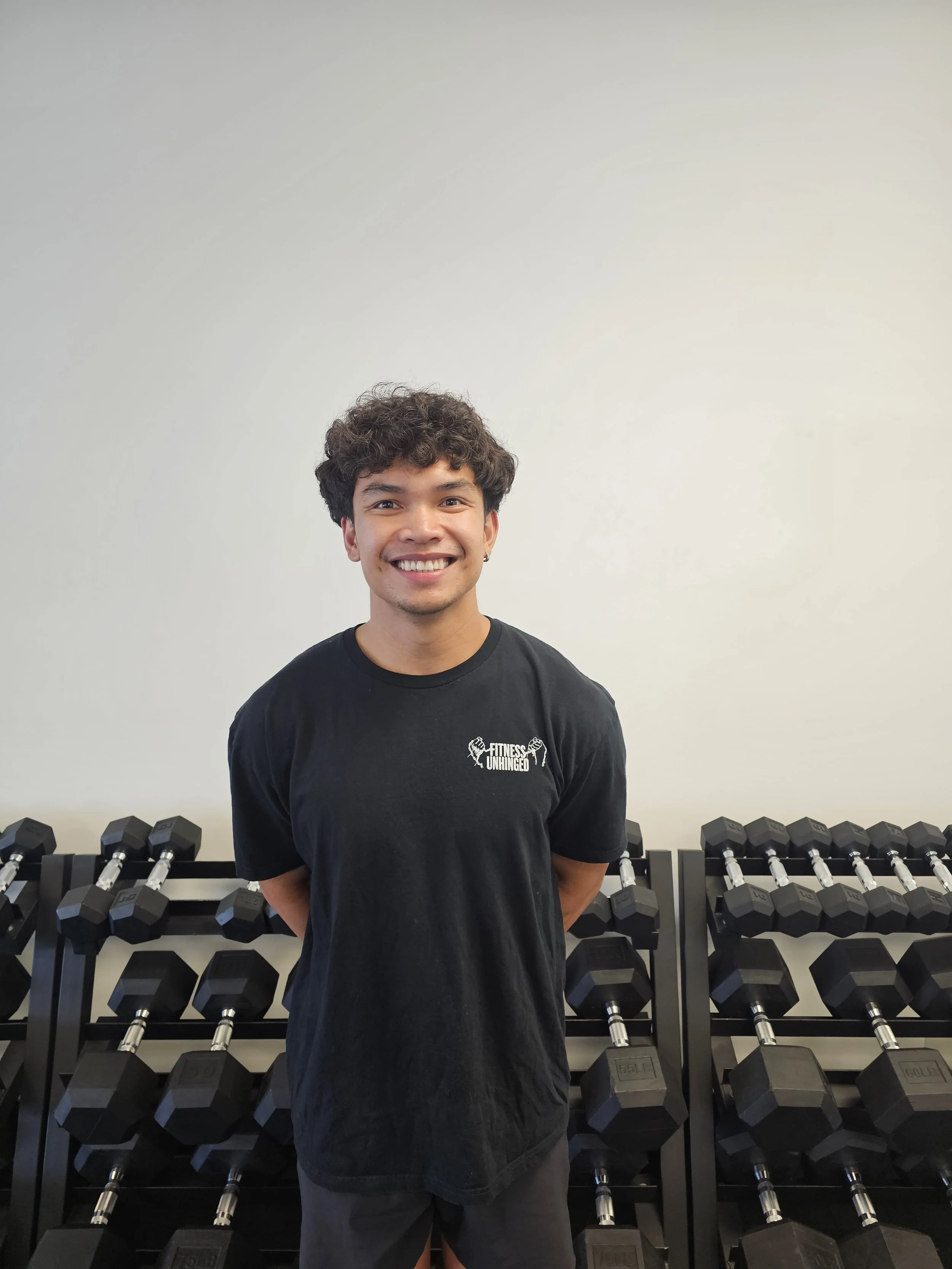 Young man with curly hair smiling in a gym, standing in front of a rack of black dumbbells against a plain light-colored wall.