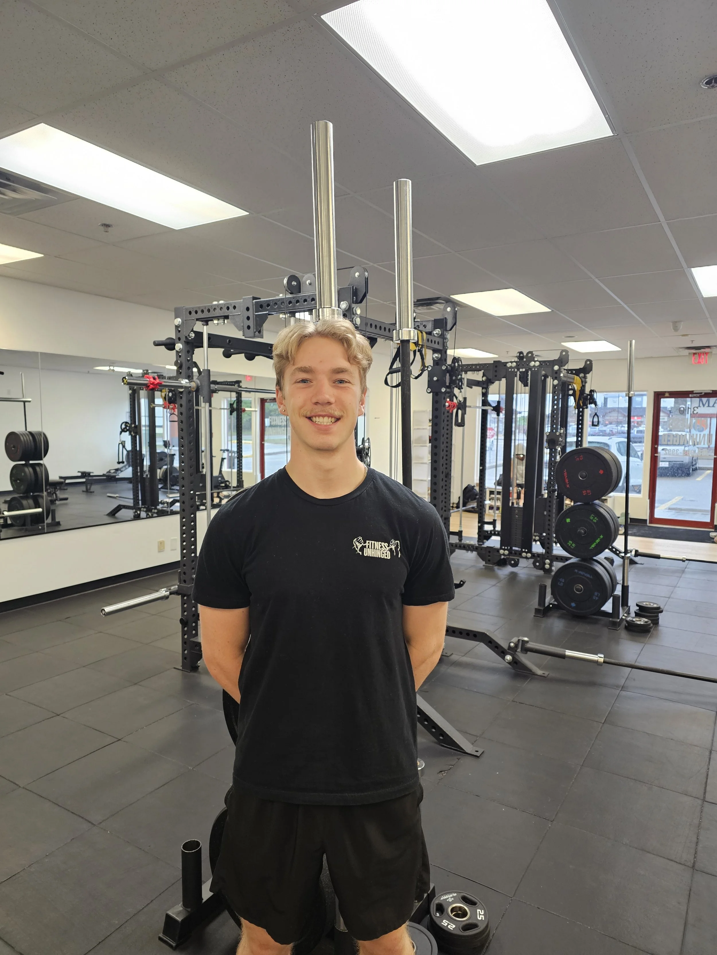 A young man with blonde hair and a mustache smiling in a gym, wearing a black shirt with 'Fitness Unhinged' written on it. The gym has various weightlifting equipment, mirrors, and large windows.