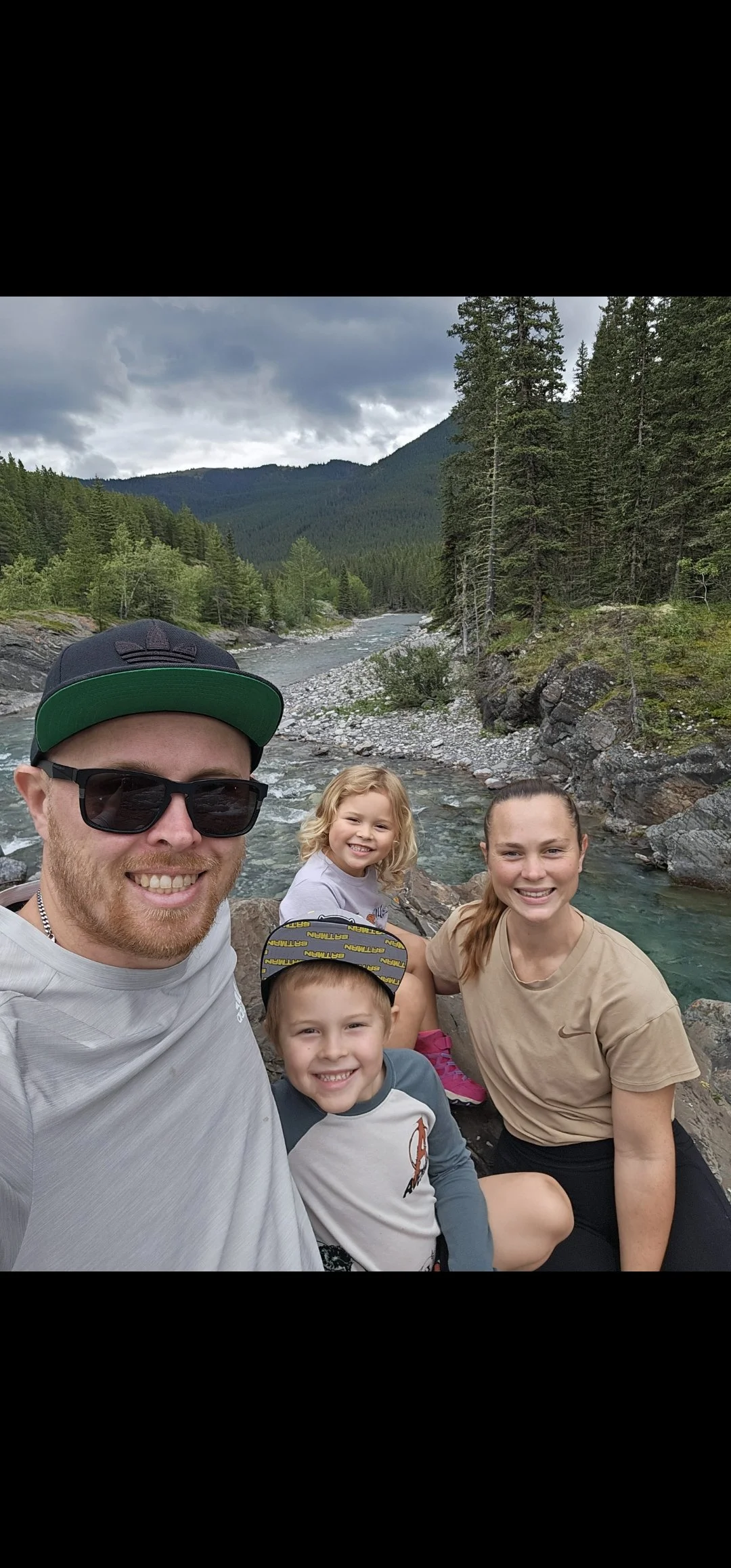 A smiling family of five, including two young girls and two boys, together outdoors by a river with forest and mountain scenery in the background.